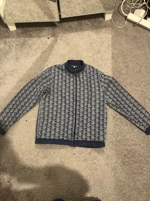 Blå tröja från Lacoste. Pris kan diskuteras. - Snygg blå bomberjacka från Lacoste med geometriskt mönster i vitt. Jackan har ribbad krage, muddar och nederkant samt dragkedja framtill med Lacoste-logga. Klassisk krokodilpatch på bröstet. Perfekt för dig som gillar streetwear och vill sticka ut.