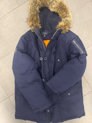Mörkblå parkajacka Polo Ralph Lauren - Parkajacka från Polo Ralph Lauren i mörkblått med fluffig fuskpäls på huvan och orange foder. Jackan har dragkedja, knappar och flera fickor, samt en Polo-patch på ärmen. Perfekt för kalla dagar med sin varma och stilrena look. Storlek 14-16 år