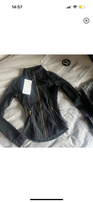 Träningsjacka lik Lululemon - Snygg svart träningsjacka som ser ut som dom från Lululemon, med guldiga dragkedjor och figurnära passform. Jackan har hög krage, långa ärmar och två fickor med dragkedja framtill. Materialet är mjukt och stretchigt syntet, perfekt för träning eller chill.🩵🩷Storlek 4 (XS)