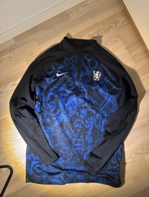 Chelsea Nike training quarter zip - Football Premier League - Snygg långärmad träningströja från Nike i slim fit med svart och blått mönster, halv dragkedja och coola detaljer. Dri-Fit material som andas och håller dig fräsch under fotboll eller annan sport. Ikonisk Nike-logga och lejonemblem på bröstet.