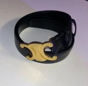 Svart läderbälte Celine Paris - Säljer ett svart bälte från Celine med ikoniskt guldspänne i form av deras Triomphe-logo. Bältet är tillverkat i slätt skinn och har en stilren, klassisk form. Perfekt för att lyfta din outfit med en lyxig detalj från Paris. Storlek 80/25mm