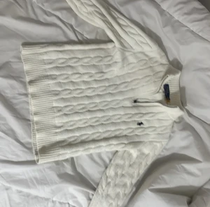 Vit kabelstickad tröja Polo Ralph Lauren - Snygg vit kabelstickad tröja från Polo Ralph Lauren med halv dragkedja och ribbad krage. Tröjan har långärmad passform och är tillverkad i mjuk bomull. Klassisk broderad logga på bröstet och breda ribbade muddar vid ärmslut och nederkant. DEN ÄR L FÖR BARN