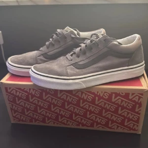 Grå Vans Old Skool sneakers  - Säljer ett par grå Vans Old Skool sneakers i storlek 37. Skorna har klassisk låg profil, snörning och vit platt sula med brun yttersula. Materialet är mocka och textil med svart sidostrip. Perfekt för dig som gillar streetstyle och skate.