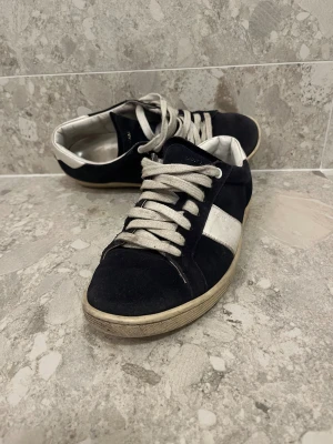Saint Laurent marinblå sneakers med vit detalj - Marinblå sneakers från Saint Laurent med vit kontrastdetalj på sidan och vit häl. Skorna har rund tå, platt sula och snörning med breda vita skosnören. Utsidan är i mocka och insidan i vitt skinn, vilket ger en lyxig känsla. Perfekt för dig som gillar stilrena och klassiska sneakers.