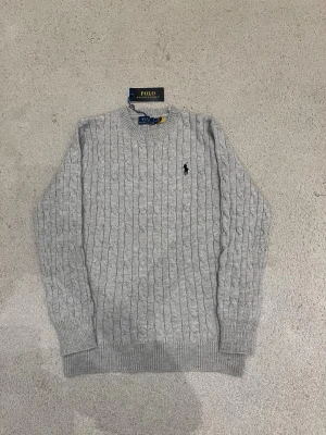 Ralph Lauren Sweatshirt  - Snygg grå kabelstickad tröja från Polo Ralph Lauren med klassisk rund hals och svart broderad logga på bröstet. Tröjan har långa ärmar och ribbade muddar vid ärmslut och nederkant. Perfekt för dig som gillar stilren och tidlös design.
