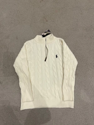 Vit stickad tröja från Polo Ralph Lauren - Säljer en vit stickad tröja från Polo Ralph Lauren med klassisk kabelstickning och half zip framtill. Tröjan har långärmad design, ribbade muddar och den ikoniska loggan broderad på bröstet. Perfekt för dig som gillar stilren och tidlös look.