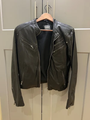 Svart skinnjacka från Livelo, stl 42 - Svart skinnjacka från Livelo med coola quiltade detaljer på axlarna och flera dragkedjor framtill. Jackan har lång ärm, ståkrage och snygga spännen bak. Är tyvärr lite sliten 