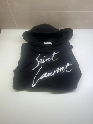 Svart Saint Laurent hoodie med silvertryck - Svart hoodie från Saint Laurent med stor huva och silverfärgat Saint Laurent-tryck på bröstet. Tröjan är i mjuk bomull och har en klassisk, loose passform. Perfekt för dig som gillar exklusiva streetwear-vibes och vill sticka ut med ett lyxigt märke.