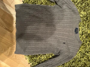 Grå kabelstickad tröja Polo Ralph Lauren - Snygg grå kabelstickad tröja från Polo Ralph Lauren i storlek xs. Klassisk design med rund hals och diskret broderad logga på bröstet. Mjuk och skön kvalitet, perfekt för lager-på-lager under höst och vinter. Långa ärmar och ribbade muddar ger en clean look. 