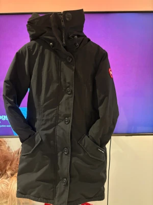 Svart parkajacka från Canada Goose - Svart parkajacka från Canada Goose med huva, stora knappar och dragkedja framtill. Jackan har två fickor med lock och snyggt broderat märke på ärmen. Materialet är slitstark polyester och jackan är fodrad för extra värme. Perfekt för kalla vinterdagar. Äkta päls. 