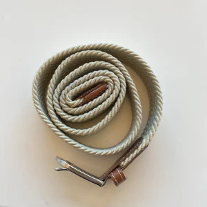 Beige flätat bälte med skinn-detaljer - Snyggt beige flätat bälte med bruna skinn-detaljer och silvrigt spänne. Bältet har en grov, rep-liknande struktur och vita sömmar på skinnet. Perfekt för dig som vill ha en stilren accessoar med lite extra karaktär.