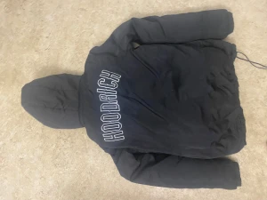 Svart pufferjacka från Hoodrich - Svart pufferjacka från Hoodrich med stor logga på ryggen och mindre logga framtill. Jackan har huva, dragkedja och långärmade ärmar. Perfekt för kalla dagar och har en clean streetwear-vibe.