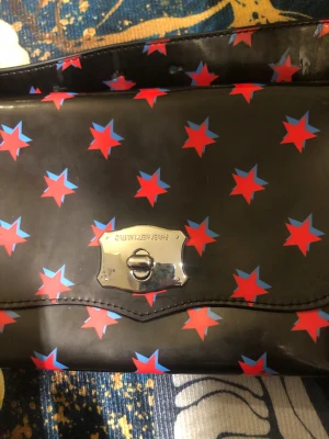 Calvin Klein axelväska med stjärnor - Cool axelväska från Calvin Klein Jeans i svart syntetmaterial med röda och blå stjärnor över hela väskan. Väskan har ett silvrigt spänne framtill och röd insida. Perfekt för dig som vill sticka ut med en unik accessoar.