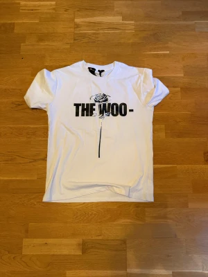 Vit t-shirt THE WOO- med rosprint - Säljer en vit t-shirt med svart tryck 'THE WOO-' och en ros på framsidan. Baksidan har en stor siffra 1 med rosdetaljer. T-shirten har rund hals och korta ärmar, perfekt för dig som gillar streetwear och snygga prints. Materialet är mjuk bomull.
