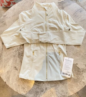  Define Jacket Nulu från Lululemon ivory - Supersnygg vit Define Jacket Nulu från Lululemon med guldfärgade dragkedjor, figurnära passform och höga krage. Jackan har tumhål, två fickor med dragkedja och är tillverkad i ett mjukt, stretchigt material som andas. Perfekt för träning eller chill.