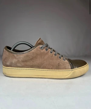 Bruna lanvin med mocka och lackad tå - Snygga bruna sneakers med ovandel i mjuk mocka och en glansig, lackad tå i mörkbrunt. Skorna har grå snörning och en beige platt sula som ger en clean look. Perfekt för dig som gillar stilrena detaljer och jordnära färger.