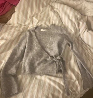 Grå tröja Gina Tricot - Mysig grå omlottopp från Gina Tricot i mjuk ullblandning. Toppen har långa ärmar, croppad passform och knyt i sidan för en snygg siluett. Perfekt att styla med jeans eller kjol för en chill look. Använd typ 1-2 gånger och inget syns på den 💖 säljer pga att den inte används 
