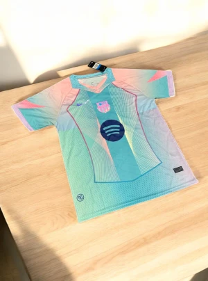 Barça x Nike pastell fotbollströja - Säljer en Barcelona fotbollströja från Nike med Spotify-logga. Tröjan har korta ärmar och är designad i pastellfärger som ljusblå, rosa och mintgrön med grafiska linjer och detaljer. Materialet är lätt och andas, perfekt för match eller träning. Snygg V-ringad krage med mönstrad kant.