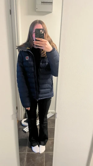 Mörkblå dunjacka från Fjällräven - Säljer en mörkblå dunjacka från Fjällräven med klassisk logga på bröstet. Jackan har quiltad design, dragkedja framtill och huva med grå insida och gul detalj. Perfekt för kalla dagar och har en normal passform.