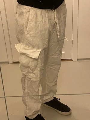Vita cargopants med snörning - Säljer ett par vita cargopants med flera stora fickor på sidorna och snörning i midjan. Byxorna har en loose passform och är tillverkade i bomull, vilket gör dem sköna och lätta. Perfekta för dig som gillar streetstyle och vill ha mycket plats för prylar.