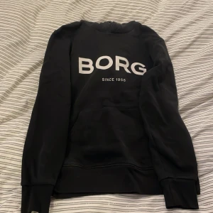 Svart hoodie från Borg - Svart hoodie från Borg med stor vit logga på bröstet och texten 'Since 1956'. Tröjan har huva, lång ärm och en klassisk magficka framtill. Perfekt för chill eller träning, enkel och stilren design.