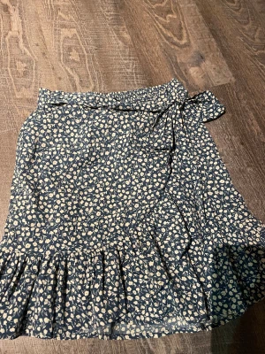 Blommig omlottkjol från ONLY - Supersöt kort omlottkjol från ONLY i mörkblått med ljusa blommor. Kjolen har volangkant och knytband i midjan som ger en extra feminin touch. Perfekt för dig som gillar romantiska och trendiga detaljer.