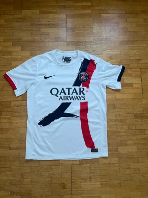 PSG vit matchtröja Nike - Paris Saint-Germain fotbollströja i vit polyester med tryckta penseldrag i marinblått och rött på framsidan. PSG-logga på bröstet, Qatar Airways sponsortryck och Nike Swoosh. Kortärmad modell med rund hals och engineered-märke nedtill.