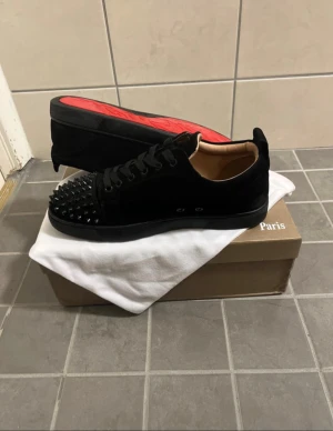 Christian Louboutin svarta sneakers med nitar - Svarta sneakers från Christian Louboutin med coola svarta nitar på tån och klassisk röd sula. Skorna har snörning och är tillverkade i mocka med en platt sula. Kommer med original dustbag och låda. Perfekt för dig som gillar edgy designerstil.