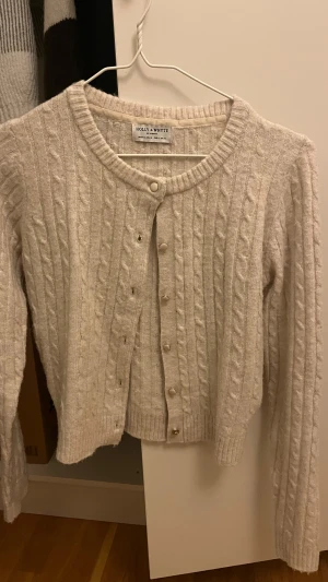 Beige kabelstickad kofta Holly & Whyte - Säljer en beige kabelstickad kofta från Holly & Whyte by Lindex i storlek XS. Koftan har rund hals, långa ärmar och snygga knappar framtill. Perfekt för lager-på-lager och chill stil. Ribbstickade muddar vid ärmslut och nederkant. Aldrig använd tyvärr