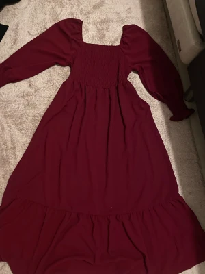 Vinröd långklänning med puffärmar - Säljer en vinröd långklänning med fyrkantig halsringning och puffiga långärmar. Klänningen har smockad överdel och volangdetalj nedtill. Materialet känns mjukt och lätt, perfekt för dig som gillar romantisk stil och vill sticka ut med färg.