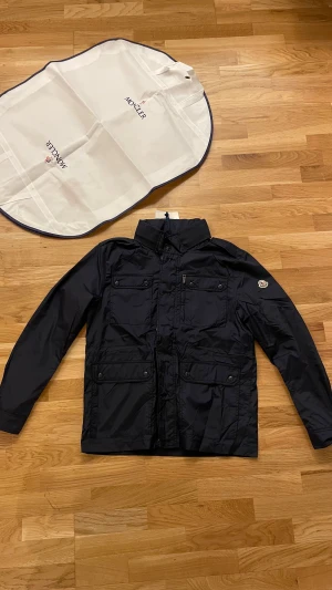 Marinblå vindjacka från Moncler - Säljer en marinblå vindjacka från Moncler med klassisk logga på ärmen. Jackan har fyra framfickor med knappar och dragkedja, hög krage och snygga detaljer. Materialet är polyester och jackan är långärmad med normal passform. Perfekt för dig som gillar stilrena och funktionella plagg.