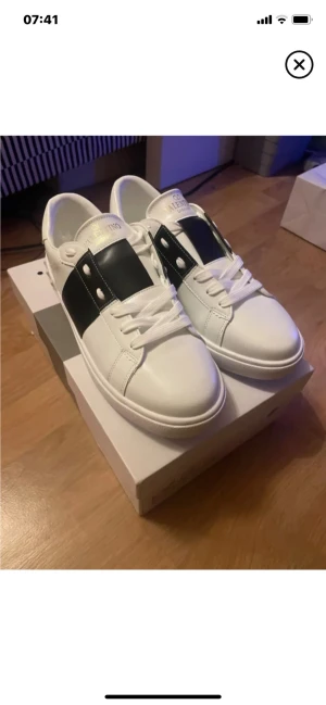 Valentino Garavani vita sneakers - Snygga vita sneakers från Valentino Garavani med bred svart rand över mitten och coola nitar på sidan. Skorna har rund tå, platt sula och klassisk snörning. Tillverkade i skinn för en lyxig känsla och stilren look. Perfekta för dig som gillar exklusiva märken och streetstyle.