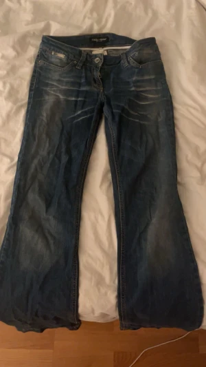 Blå bootcut jeans från Dolce & Gabbana - Snygga blå jeans från Dolce & Gabbana med bootcut passform och klassisk femficksdesign. Jeansen har slitningar och tvättade detaljer framtill, samt metallknapp och logotyp på fickan. Tillverkade i jeansmaterial med låg midja.
