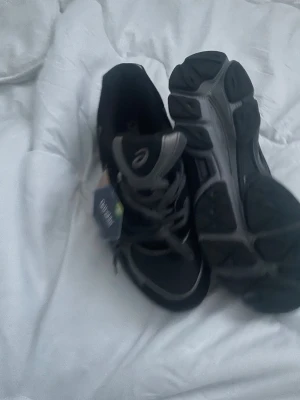  Asics Skor - Säljer ett par svarta Asics sneakers med grå detaljer och Ortholite komfortsula. Skorna har snörning och en sportig design med rund tå och platt sula. Materialet är syntet och mesh för bra andningsförmåga. Perfekt för dig som gillar stilrena och funktionella träningsskor.