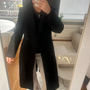 Svart kappa med bälte och bred krage - Snygg svart kappa med bred krage och knytbälte i midjan. Kappan har en klassisk, rak passform och stängs med en knapp framtill. Perfekt för dig som gillar stilrena och tidlösa plagg. Materialet är tjockt och känns varmt för kyliga dagar. Köpt på Mango förra året för 2199kr. Xs men funkar även till mig som egentligen är S