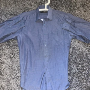 Blå skjorta från Yves Saint Laurent - Klassisk blå skjorta från Yves Saint Laurent med diskret YSL-broderi på bröstfickan. Skjortan har lång ärm, knappar framtill och en stilren krage. Storleken är L!