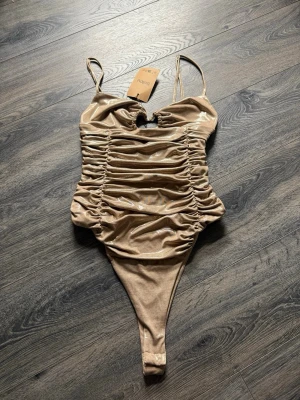 Glittrig beige body från Sellpy, men från början från Zara - Säljer en glittrig beige body från Sellpy/Zara med tunna axelband och rynkade detaljer framtill. Materialet har en skimrande finish som ger en extra touch.