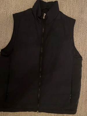 Svart dunväst från Oscar Jacobson - Säljer en marinblå dunväst från Oscar Jacobson med hög krage och hel dragkedja framtill. Västen har en stilren och enkel design, perfekt att slänga över en hoodie eller tröja. Materialet är mjukt och lätt, vilket gör den smidig att bära under kyligare dagar.