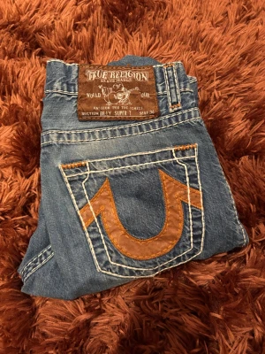 True Religion blå jeans med kontrastsömmar - Säljer ett par klassiska blå jeans från True Religion, modell Billy Super T. Jeansen har tydliga kontrastsömmar i vitt och orange samt stora bakfickor med ikonisk broderad logga i brunt. Tillverkade i slitstark denim med snygg tvätt och detaljerade nitar❤️❤️