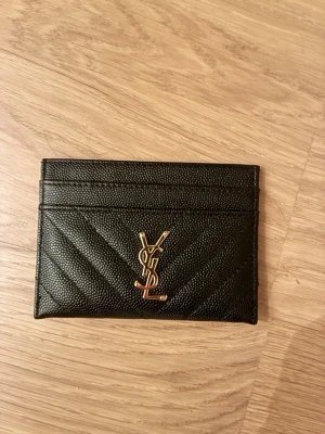 Svart YSL plånbok  - Säljer en svart plånbok från YSL med guldfärgad logga framtill. Plånboken har ett stilrent quiltat mönster och flera fack för kort. Perfekt accessoar för dig som gillar lyxiga detaljer. 