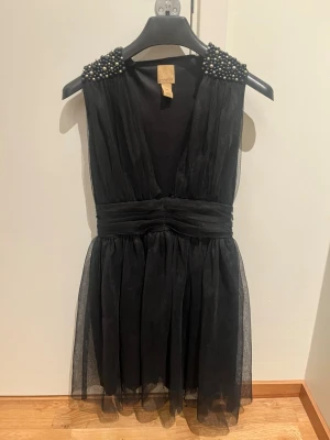 Svart kort klänning med pärldetaljer - Supersnygg svart kort klänning från Jeane Blush med djup v-ringning och markerad midja. Axelpartiet är dekorerat med svarta och silvriga pärlor som ger en lyxig känsla. Klänningen är ärmlös och har ett skirt, lätt tyg som ger ett elegant fall.