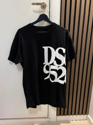 Svart DSQUARED2 t-shirt med tryck - Säljer en svart t-shirt från DSQUARED2 med stort vitt tryck 'DS 952' på framsidan och liten logga i nacken. Klassisk rund hals och korta ärmar. Tillverkad i mjuk bomull för skön känsla och snygg passform. Kom dm för eventuella frågor 🧐pris kan diskuteras 😇💯