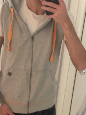 Grå ärmlös hoodie med orange snören - Säljer en grå ärmlös hoodie med dragkedja framtill och huva. Tröjan har orange snören i huvan och två stora fickor på magen. Perfekt för lager på lager och chill stil. Materialet känns mjukt och bekvämt, troligen bomull eller bomullsblandning. Vid intresse skriv för mer info! ABS! Pris går att diskutera!