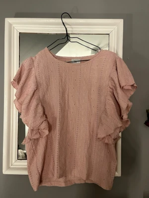 Rosa volangblus från Isolde - Supersöt ljusrosa blus från Isolde med voluminösa volangärmar och diskret strukturmönster. Blusen har rund halsringning och ett subtilt geometriskt mönster i tyget som ger en extra touch. 