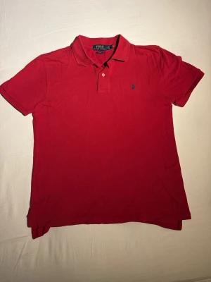 Röd pikétröja Polo Ralph Lauren - Klassisk röd pikétröja från Polo Ralph Lauren i slim fit-modell. Tröjan har korta ärmar, krage med två knappar och den ikoniska blå broderade loggan på bröstet. Tillverkad i mjuk bomull som andas och passar perfekt till jeans eller shorts.