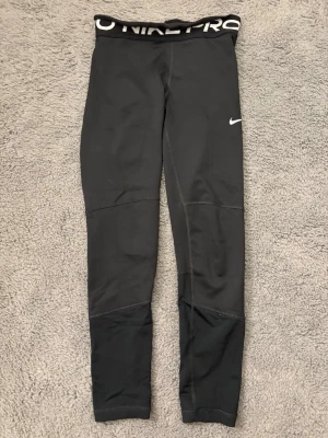 Svarta Nike Pro tights 🏋️‍♀️ - Säljer ett par svarta Nike Pro tights med vit Nike-logga på låret och bred resår i midjan med Nike Pro-tryck. Tillverkade i stretchigt syntetmaterial som passar perfekt för träning och sportiga aktiviteter.