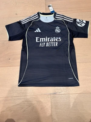 Real Madrid tröja – Mbappé #10 -  Svart Real Madrid-tröja från Adidas med Kylian Mbappé #10 på ryggen. Stilren design med Emirates Fly Better-loggan och Real Madrid-emblemet. AEROREADY-material som andas bra och är skönt att bära.  📌 Detaljer: 	•	Lag: Real Madrid 	•	Spelare: Mbappé 	•	Nummer: 10 	•	Märke: Adidas 	•	Färg: Svart 	•	Passform: Normal 	•	Skick: Mycket bra (inga hål eller fläckar)  Perfekt för match, träning eller casual outfit. Skickas snabbt 🚚 Hör av dig vid frågor! 