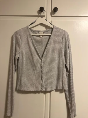 Grå ribbad kofta från H&M - Säljer en ljusgrå ribbad kofta från H&M i storlek M. Koftan har v-ringning, långa ärmar och knappar framtill. Perfekt att slänga över en topp eller bära stängd för en enkel och clean look.