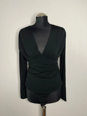 Svart omlott body från H&M - Säljer en svart body från H&M med omlott och djup v-ringning. Långa ärmar och snygg drapering över magen ger en riktigt schysst siluett. Perfekt till höga jeans eller kjol för en clean look. Använd en gång… 