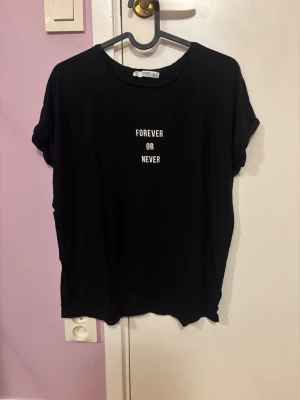 Svart t-shirt från Mango med tryck - Svart t-shirt från Mango Basics med vit text 'FOREVER OR NEVER' på bröstet. Klassisk rund halsringning och korta ärmar med lätt upprullad kant. Tillverkad i mjuk bomull för en skön och avslappnad look.